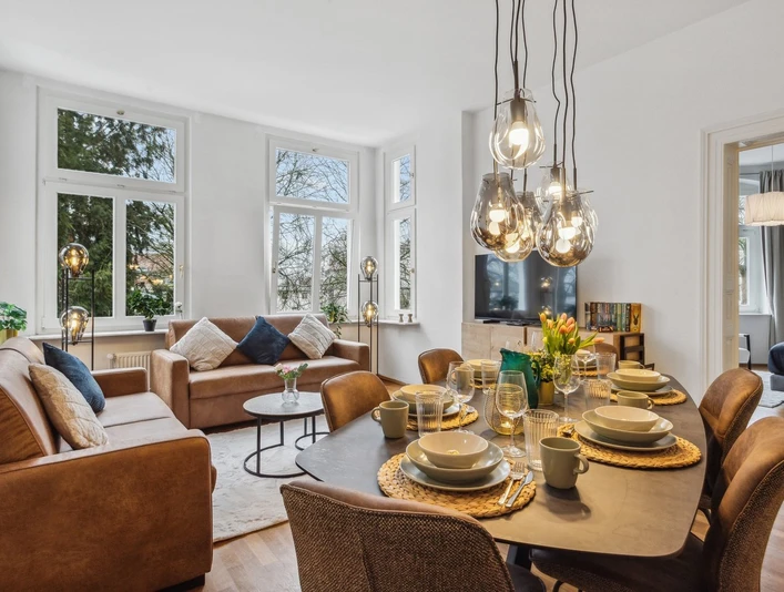 WohnLiebe_Augustaplatz2.jpeg Modernes Wohnzimmer mit großen Fenstern, bequemen Sofas, Esstisch und stilvollen Hängeleuchten.