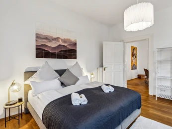 WohnLiebe_Augustaplatz3.jpeg Helles Schlafzimmer mit einem großen Bett, moderner Kunst an den Wänden und hölzernem Boden.