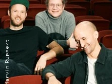 Drei lächelnde Männer sitzen entspannt in roten Kinosesseln, in lockerer Freizeitkleidung.