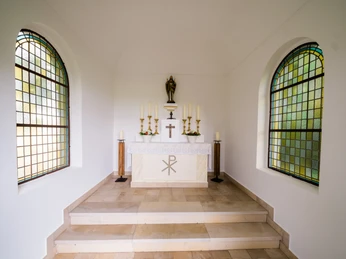 Innenansicht einer kleinen Kapelle mit Altar, verziert mit Kerzen, einem Kreuz und bunten Fenstern.