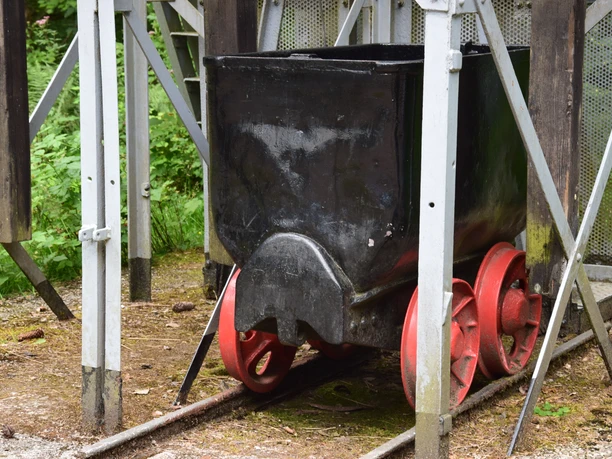 Lore am Franziskaschacht <p>Ein schwarzer Erz- oder Kohlewagen mit roten Rädern steht auf Schienen in einem industriellen Umfeld.</p>