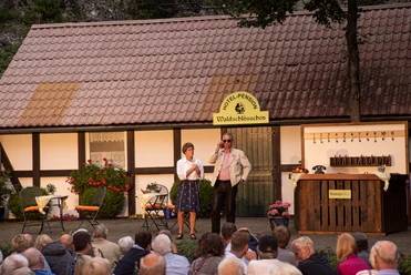 Open-Air-Theateraufführung vor einem Fachwerkhaus, Publikum sieht zwei Schauspielern in Kostümen zu.