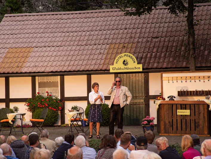 Open-Air-Theateraufführung vor einem Fachwerkhaus, Publikum sieht zwei Schauspielern in Kostümen zu.