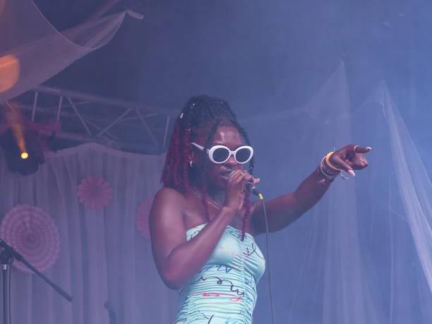 Koelnchella_23.jpg Sängerin mit Mikrofon performt in farbigem Outfit auf einer nebeligen Festivalbühne.