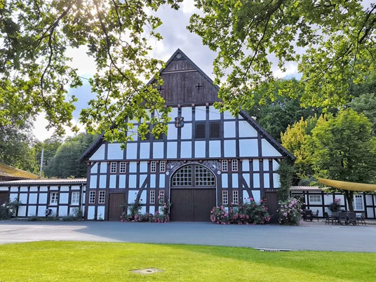 Historisches Fachwerkgebäude in ländlicher Umgebung, umgeben von Bäumen und blühenden Sträuchern.