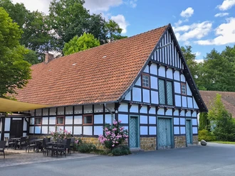 Fachwerkhaus mit großen Dach, Terrasse mit Sitzgelegenheiten, blühende Sträucher und Bäume drumherum.