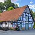 Fachwerkhaus mit großen Dach, Terrasse mit Sitzgelegenheiten, blühende Sträucher und Bäume drumherum.