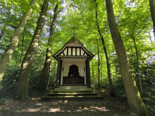 Eine kleine, weiße Kapelle mit Fachwerkelementen, umgeben von dichtem, grünem Wald.