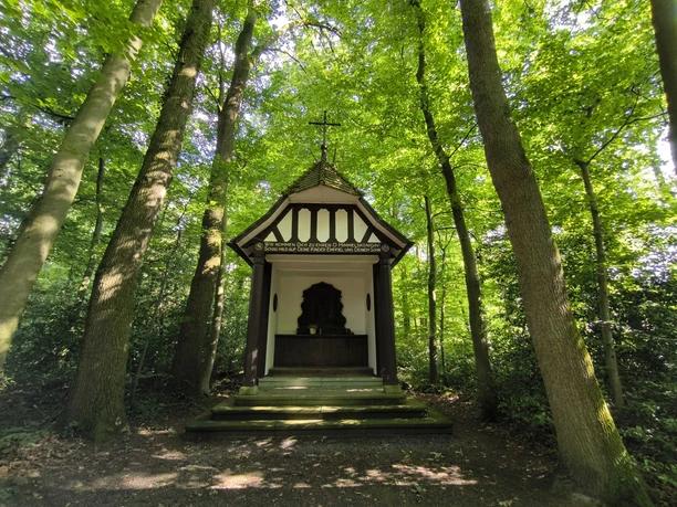 Eine kleine, weiße Kapelle mit Fachwerkelementen, umgeben von dichtem, grünem Wald.