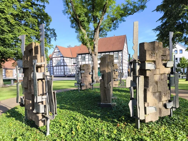 Mahnmal aus Steinblöcken im Schlossgarten Hövelhof, umgeben von Fachwerkhäusern und Bäumen.
