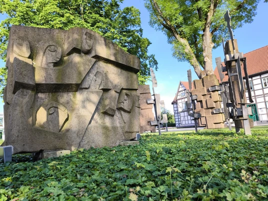 Steinskulpturen in einem Park, umgeben von grüner Vegetation. Fachwerkhaus im Hintergrund.