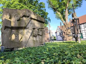 Mahnmal im Schlossgarten Hövelhof Steinskulpturen in einem Park, umgeben von grüner Vegetation. Fachwerkhaus im Hintergrund.