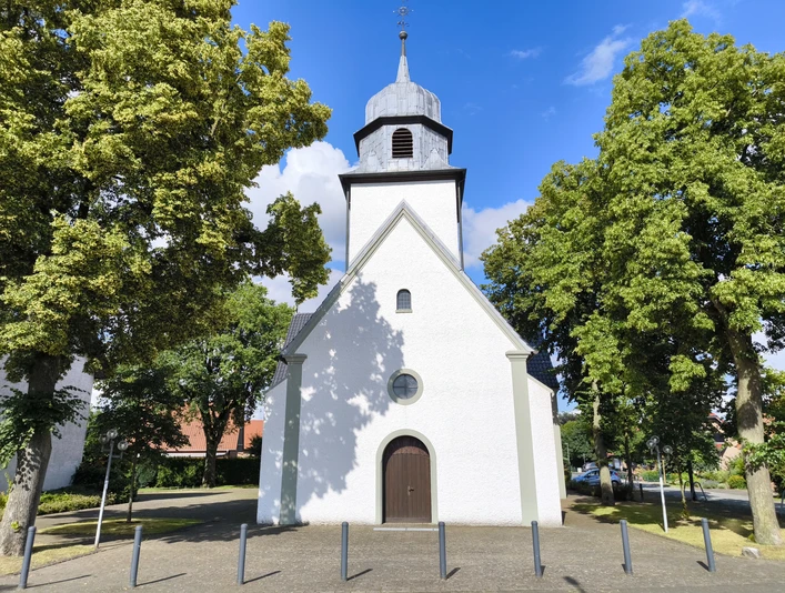 Weiße Kirche mit Holztür und Zwiebelturm, umrahmt von Bäumen und blauem Himmel.