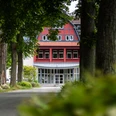 Rotes Gebäude mit Fachwerk und Glasfront in Hövelhof, umgeben von großen Bäumen und grüner Natur.