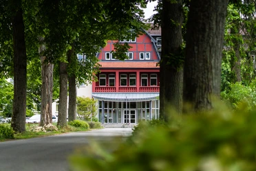 Salvator Kolleg Hövelhof Rotes Gebäude mit Fachwerk und Glasfront in Hövelhof, umgeben von großen Bäumen und grüner Natur.