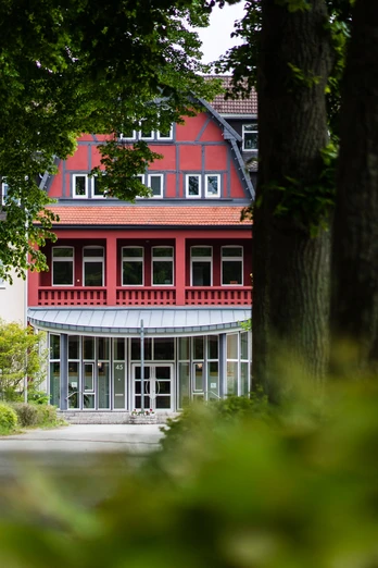 Salvator Kolleg Hövelhof Rotes Gebäude mit Fachwerk und Glasfront in Hövelhof, umgeben von großen Bäumen und grüner Natur.