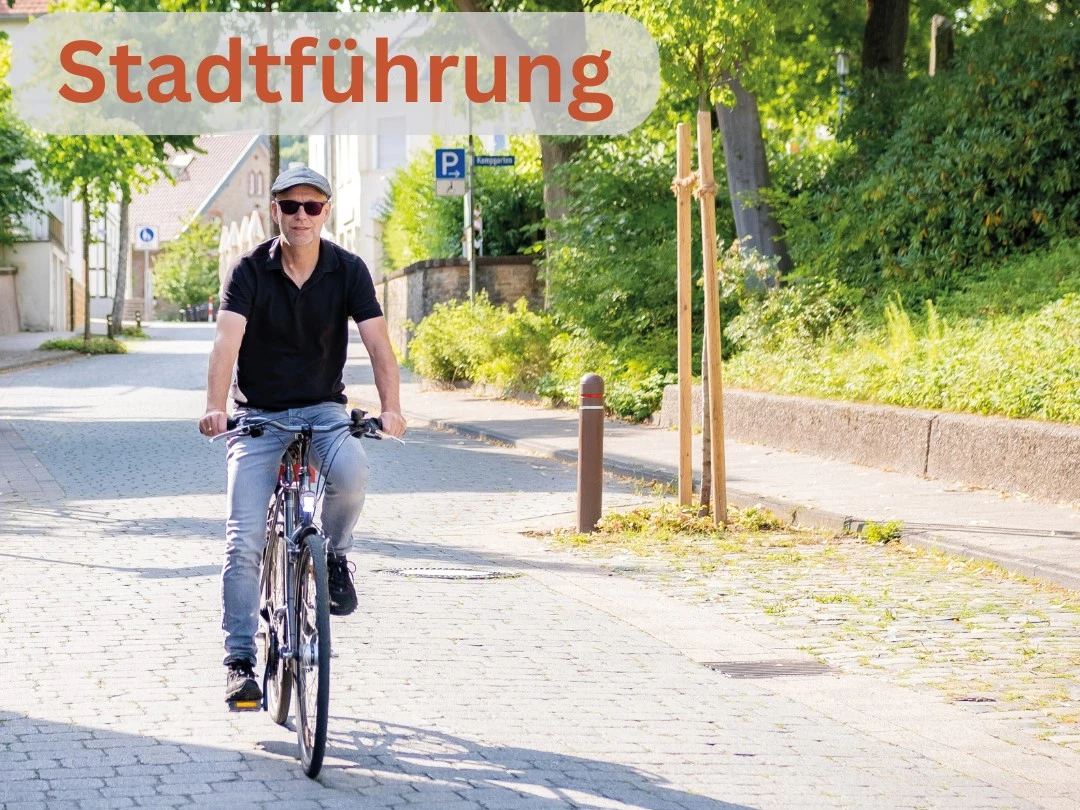Stadtführung Rädel.jpg Ein Mann fährt bei Sonnenschein auf einem blauen Fahrrad eine schattige Kopfsteinpflasterstraße entlang.