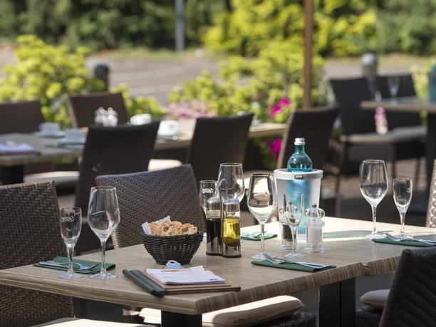 Außenterrasse im Hotel am Wald in Monheim am Rhein Gedeckte Tische auf einer sonnigen Hotelterrasse mit grüner Natur und bunten Pflanzen im Hintergrund.
