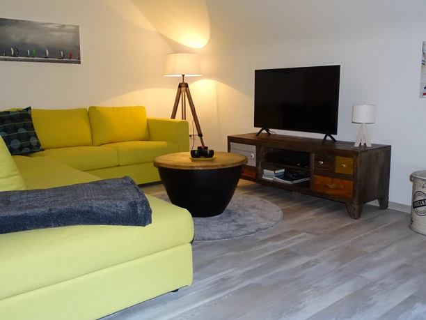 Wohnbereich in der Panorma Ferienwohnung am neuen Haus Heller Wohnbereich mit gelbem Ecksofa, Flachbildfernseher, stilvoller Beleuchtung und maritimer Dekoration.