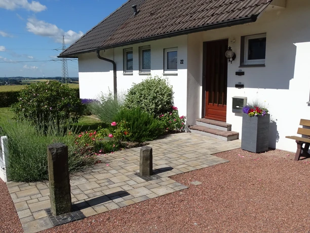 Panorma Ferienwohnung am neuen Haus Das Bild zeigt den Eingangsbereich einer Ferienwohnung mit einem gepflegten Garten und einer Holzbank.