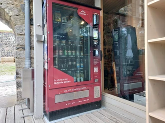 Proviantomat in Stolpen Roter Verkaufsautomat mit Snacks und Getränken, aufgestellt neben einer Steinmauer und einem Holzregal.