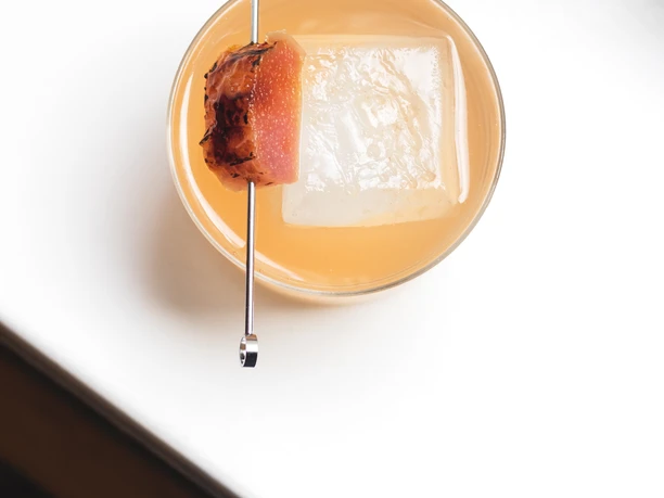Lokal (c) CSTHEROOM (1).jpg Getränk mit einem großen Eiswürfel und einer Frucht, die auf einem Spießaufgespießt ist.