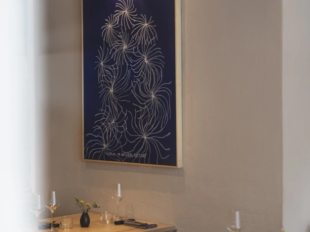 Wand mit einem Bild und kleiner Lampe darüber. Vor der Wand steh ein Tisch mit mehreren Stühlen.