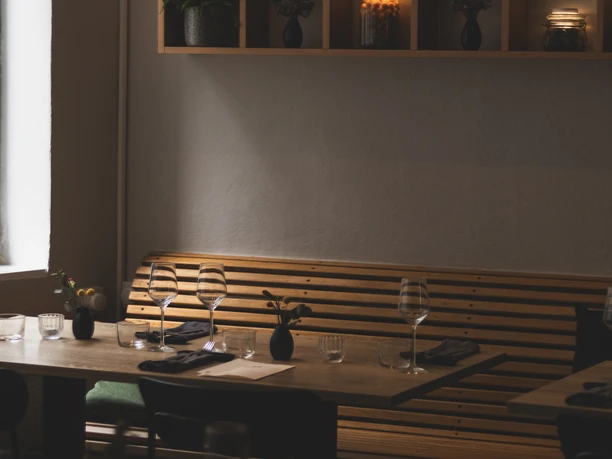 Lokal (c) CSTHEROOM (10).jpg Gedeckter Tisch mit Sitzbank an der Wand und einem Regal darüber, in dem Topfpflanzen und Lichter stehen.