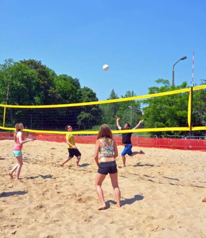 leipziger-stadthafen-beachvolleyball-foto-andreas-schmidt-800x530.jpg Menschen spielen Beachvolleyball auf einem Sandplatz unter blauem Himmel, umgeben von Bäumen.