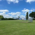 1. Sportplatz Wiefelstede Am Breeden.jpg Weitläufiger grüner Fußballplatz in Wiefelstede mit Toren, umgeben von Bäumen und blauem Himmel.