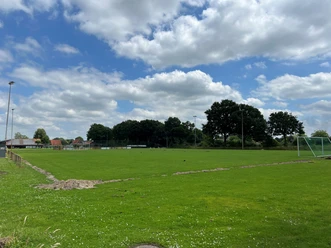 Sportplatz Metjendorf.jpg Ein weitläufiger, grüner Sportplatz in Metjendorf unter einem teils bewölkten Himmel. Im Hintergrund sind Bäume und vereinzelt Gebäude sichtbar.