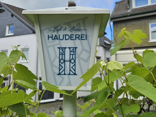 Hauderei Café & Bistro Weiße Laterne mit der Aufschrift "Café & Bistro Hauderei" inmitten grüner Pflanzen, vor Häusern.
