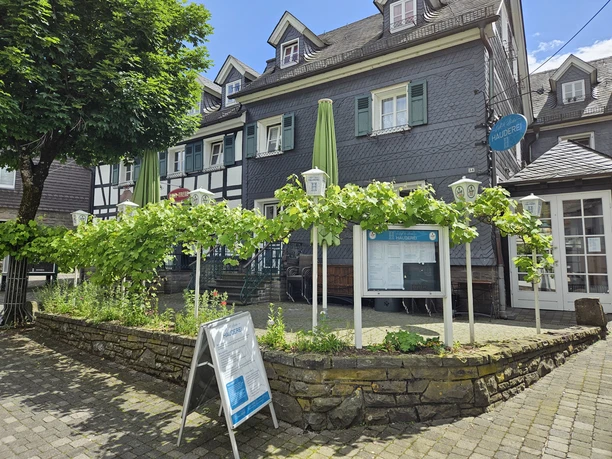 Hauderei Café & Bistro Historisches Fachwerkgebäude mit Außenterrasse, umgeben von Weinreben und Infotafeln unter blauem Himmel.