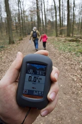Geocaching GPS-Gerät zeigt Wanderroute im Wald, dahinter laufen zwei Personen auf einem Laubweg.