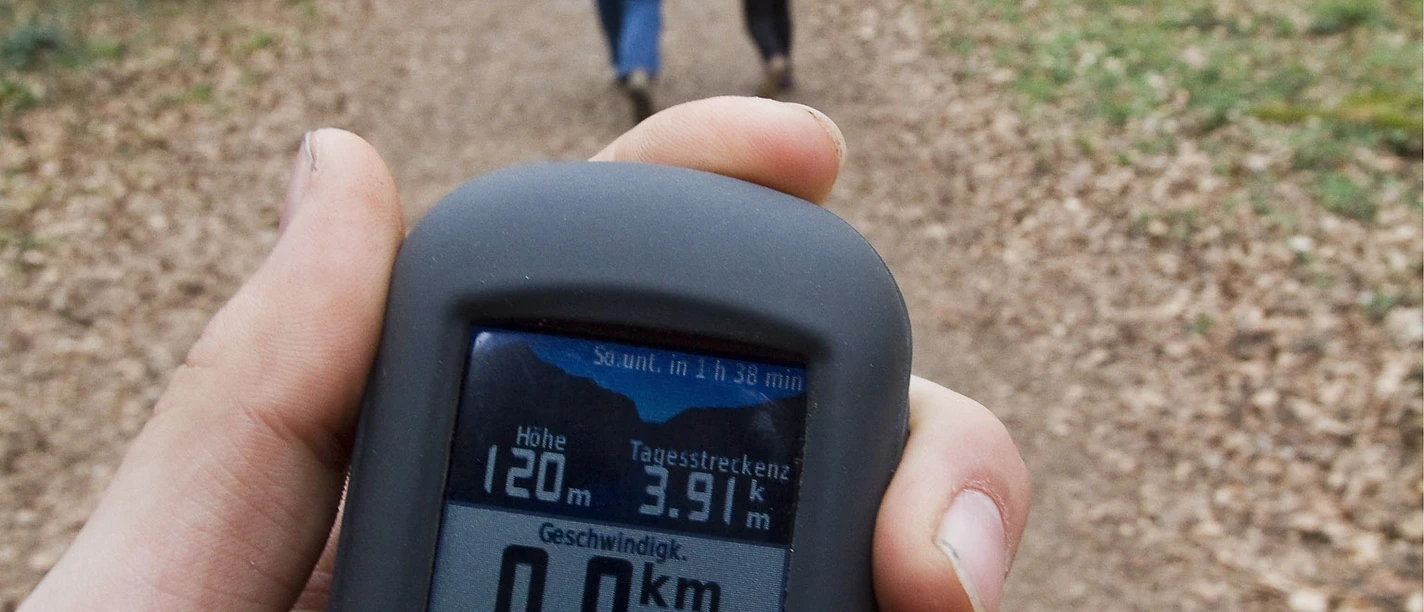 Geocaching GPS-Gerät zeigt Wanderroute im Wald, dahinter laufen zwei Personen auf einem Laubweg.