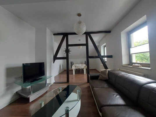 Wohnraum in der Ferienwohnung "An der alten Post" in Heiligenhaus. Wohnbereich mit Ledersofa, Glastisch, TV, Holzbalken und Essbereich in heller Ferienwohnung.