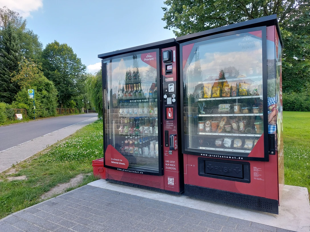 Proviantomat in Ostrau Roter Verkaufsautomat mit Snacks und Getränken, aufgestellt neben einer Landstraße, umgeben von grünen Bäumen und Wiese unter blauem Himmel.