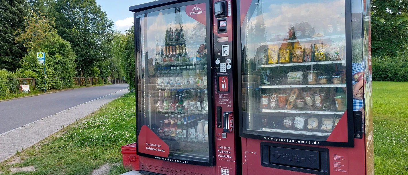 Proviantomat in Ostrau Roter Verkaufsautomat mit Snacks und Getränken, aufgestellt neben einer Landstraße, umgeben von grünen Bäumen und Wiese unter blauem Himmel.