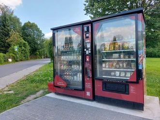 Verkoopautomaat voor levensmiddelen in Ostrava Rode automaat met snacks en drankjes, opgesteld naast een landweggetje, omringd door groene bomen en weiden onder een blauwe hemel.