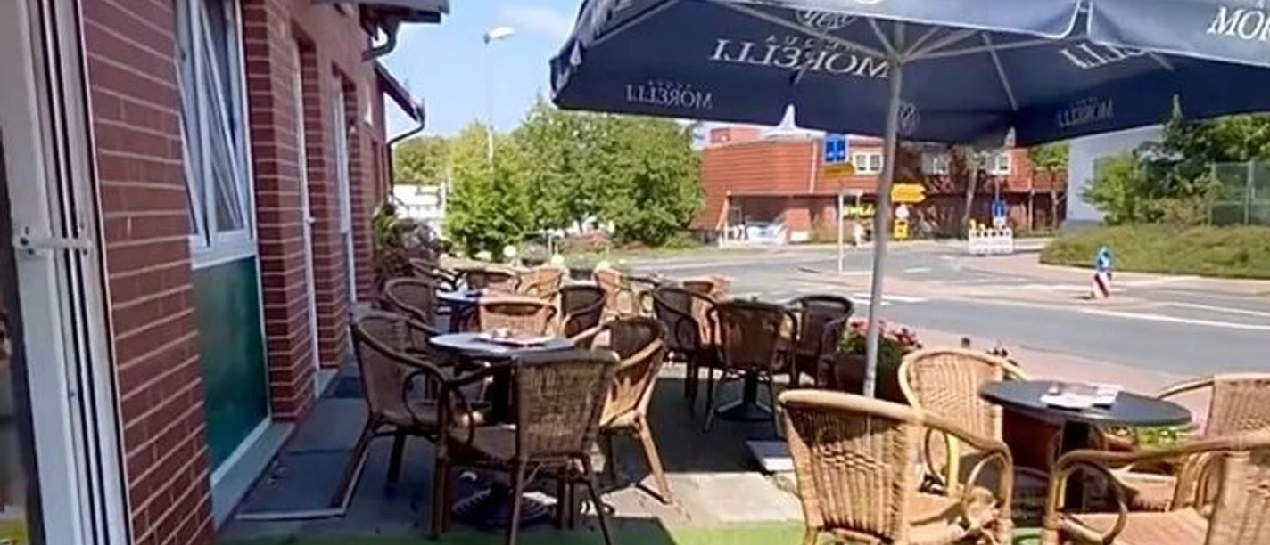 cortina.jpg Das Bild zeigt die Außenterrasse mit mehreren Tischen und Stühlen unter einem großen Sonnenschirm. Im Hintergrund ist eine Straße mit einigen Gebäuden und Bäumen zu sehen, während links eine Backsteinfassade mit einer Tür zu erkennen ist.