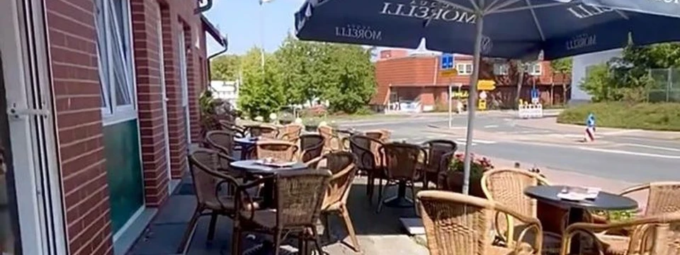 cortina.jpg Das Bild zeigt die Außenterrasse mit mehreren Tischen und Stühlen unter einem großen Sonnenschirm. Im Hintergrund ist eine Straße mit einigen Gebäuden und Bäumen zu sehen, während links eine Backsteinfassade mit einer Tür zu erkennen ist.