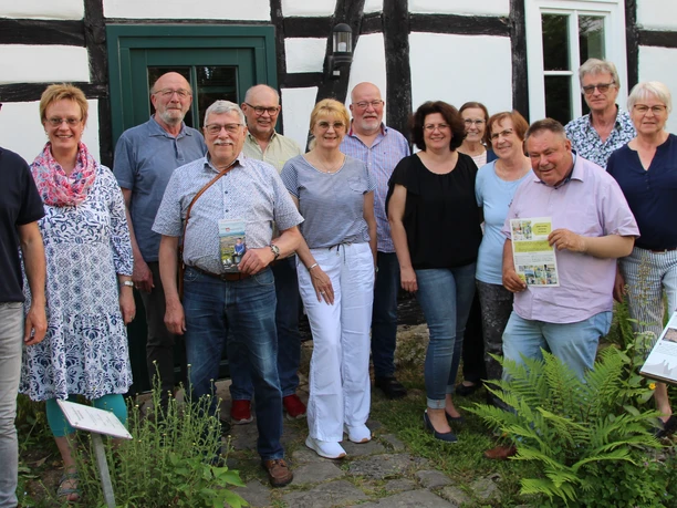 Gruppe glücklicher Menschen posiert vor einem Fachwerkhaus, umgeben von grünen Pflanzen.