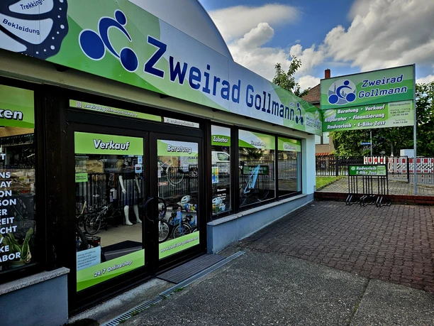 Eingangsbereich Zweirad Gollmann Fahrradgeschäft "Zweirad Gollmann" mit grüner und blauer Beschilderung, Schaufenster mit Fahrrädern und Zubehör, sonnige Atmosphäre.