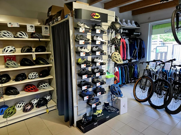 Salesroom with various models Bicycle store with shelves full of helmets and shoes, colorful jackets hanging next to them; several bicycles on the right.