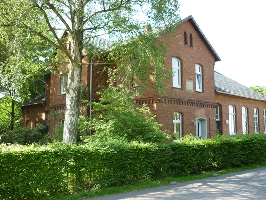 Ehemalige Backsteinschule im Grünen, umgeben von Bäumen und gepflegter Hecke, mit Satteldach.