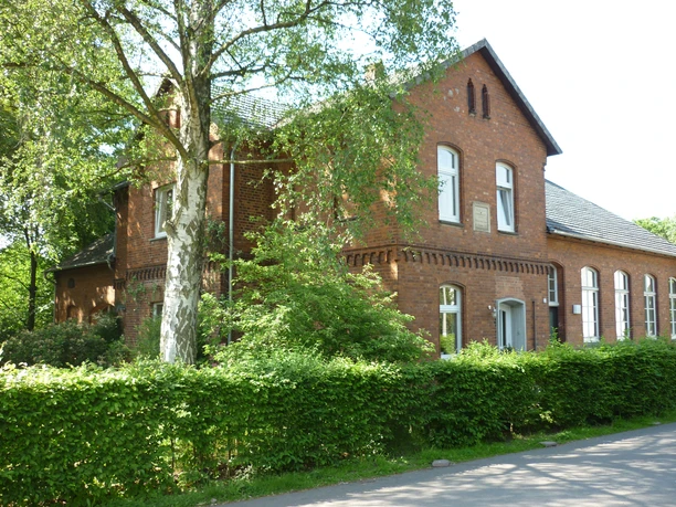 Ehemalige Backsteinschule im Grünen, umgeben von Bäumen und gepflegter Hecke, mit Satteldach.