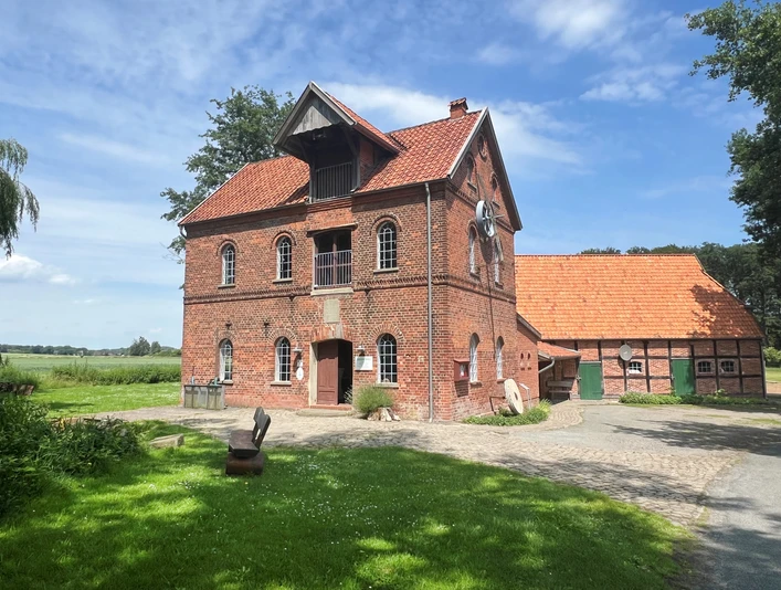 Noltesche Mühle Bruchhausen-Vilsen (2).jpg Historische Backsteinmühle mit zwei Etagen, umgeben von grünem Rasen, Bäumen und einem klaren Himmel.