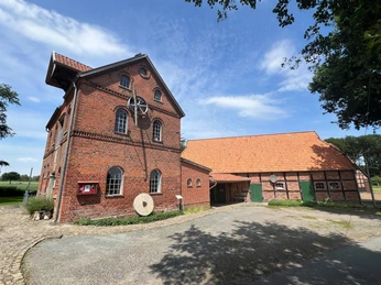 Noltesche Mühle Bruchhausen-Vilsen (3).jpg Historische rote Backsteinmühle mit Rad und Nebengebäude, umgeben von Bäumen und freiem Himmel.