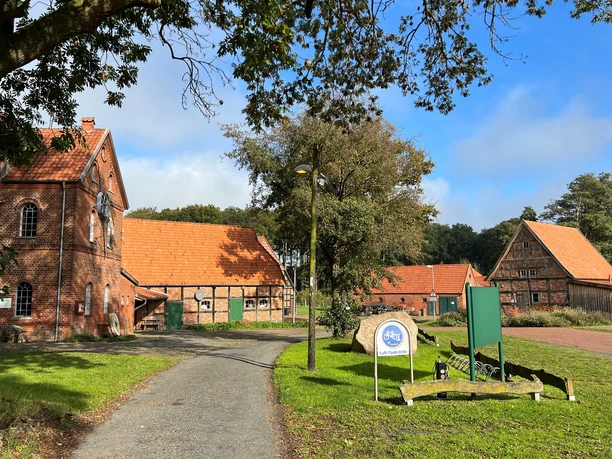 Noltesche Mühle Bruchhausen-Vilsen (5).jpg Historische Backsteinmühle in Bruchhausen-Vilsen mit Nebengebäuden und Sitzbereich unter Bäumen.