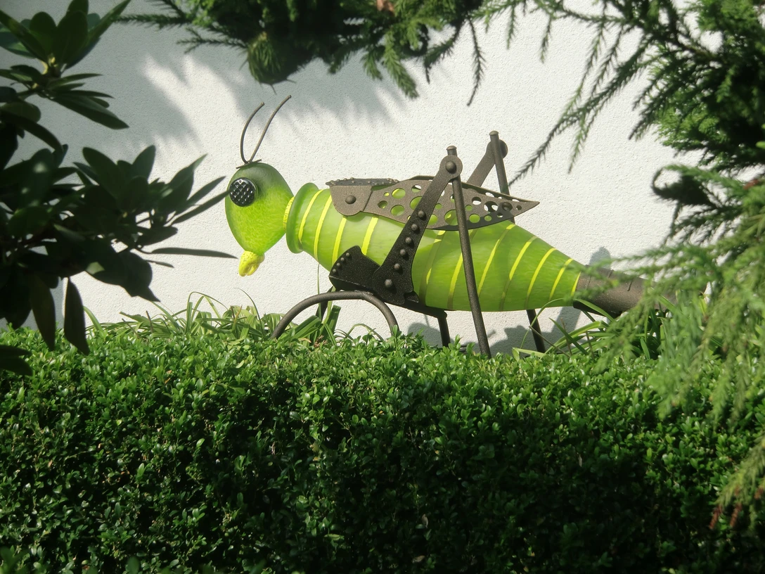 Grüne Grashüpfer-Skulptur aus Metall, umgeben von dichten Sträuchern und Blättern im Sonnenlicht.